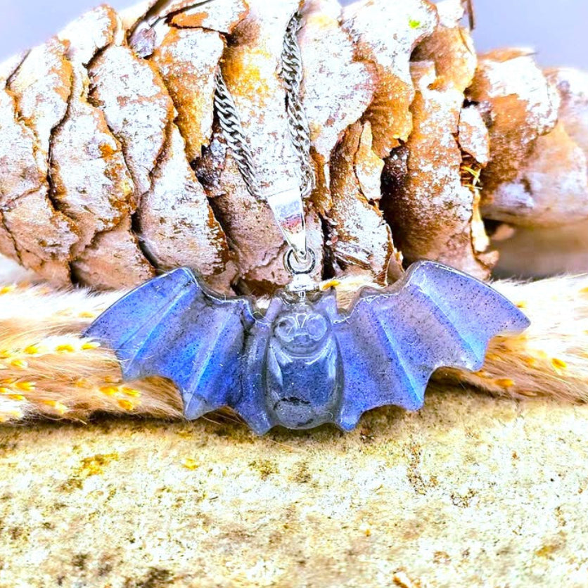 A blue natural labradorite carved bat pendant necklace with a silver-toned chain, displayed on a textured surface in front of a pinecone.-My Aura Gemstones