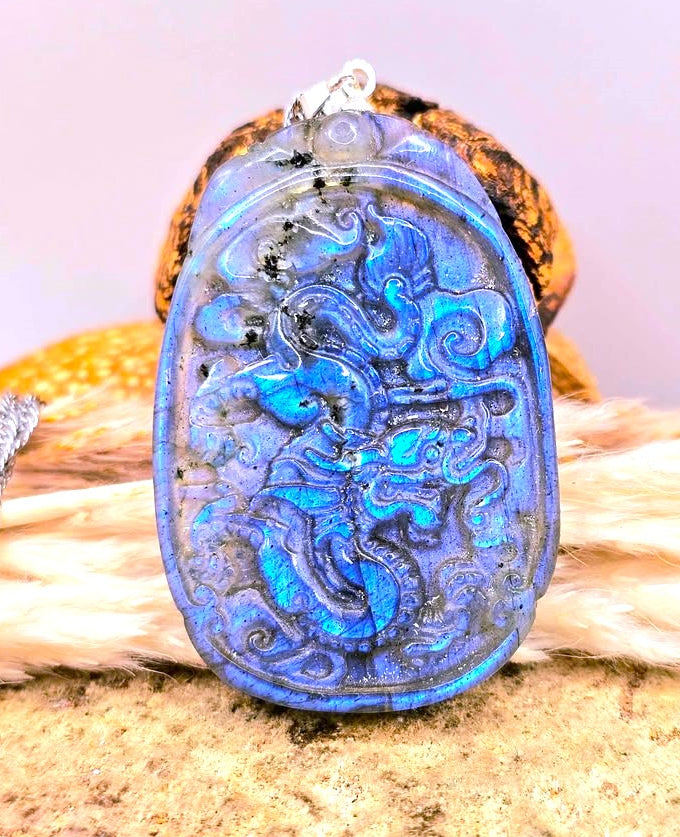 A carved blue stone pendant with an intricate dragon design, resting on a textured surface with a small silver chain visible on the left.-My Aura Gemstones