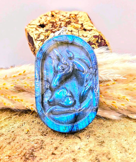 An oval cabochon pendant featuring a carved blue Unicorn motif, set in a vivid iridescent blue border. The pendant is displayed on a textured cork block and dried grass against a soft, light background.-My Aura Gemstones