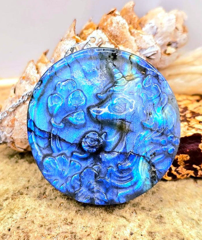 A round blue labradorite  pendant with a carved unicorn design, resting on a textured sandy surface with dried seashells behind it.- My Aura Gemstones