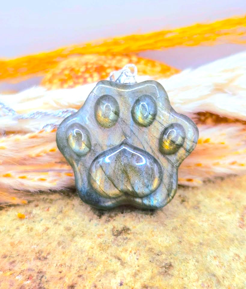 A paw-shaped pendant made of polished labradorite stone, resting on a textured surface with a blurred background of orange and white.-My Aura Gemstones