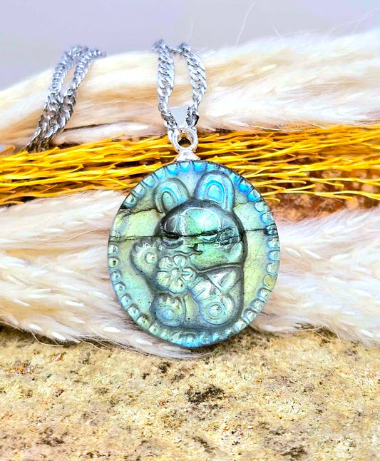 A silver chain necklace with a round pendant featuring a stylized rabbit design in iridescent blue, green, and gold tones, displayed on a textured surface with faux fur and dried grass.-My Aura Gemstones