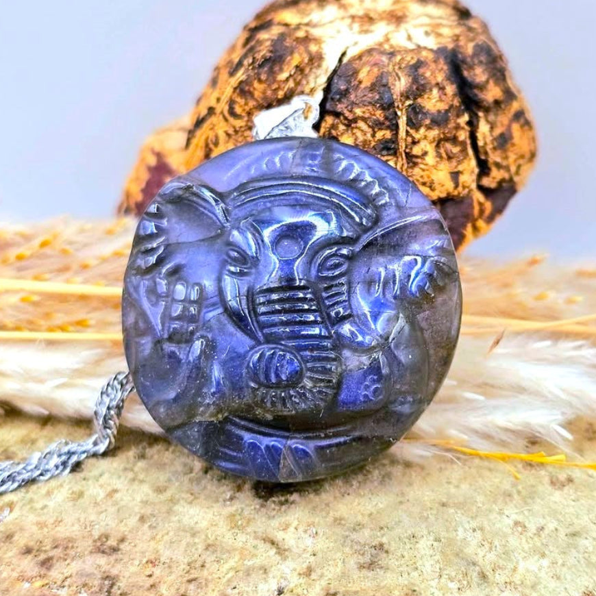 A round shape natural labradorite carved Ganesha elephant god pendant necklace in deep resting on a textured stone surface with dried grass.-My Aura Gemstones
