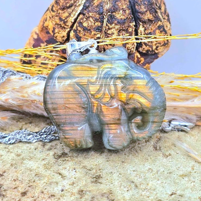 A carved elephant pendant with a smoky gray and gold striped pattern, resting on a textured surface with a feather and dried flower in the background.-My Aura Gemstones