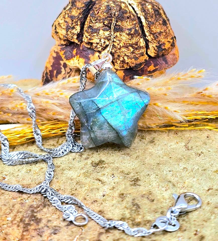 A faceted star-shaped pendant made of iridescent labradorite stone with a silver chain necklace, styled on a textured surface with dried wheat and a decorative object in the background.-My Aura Gemstones