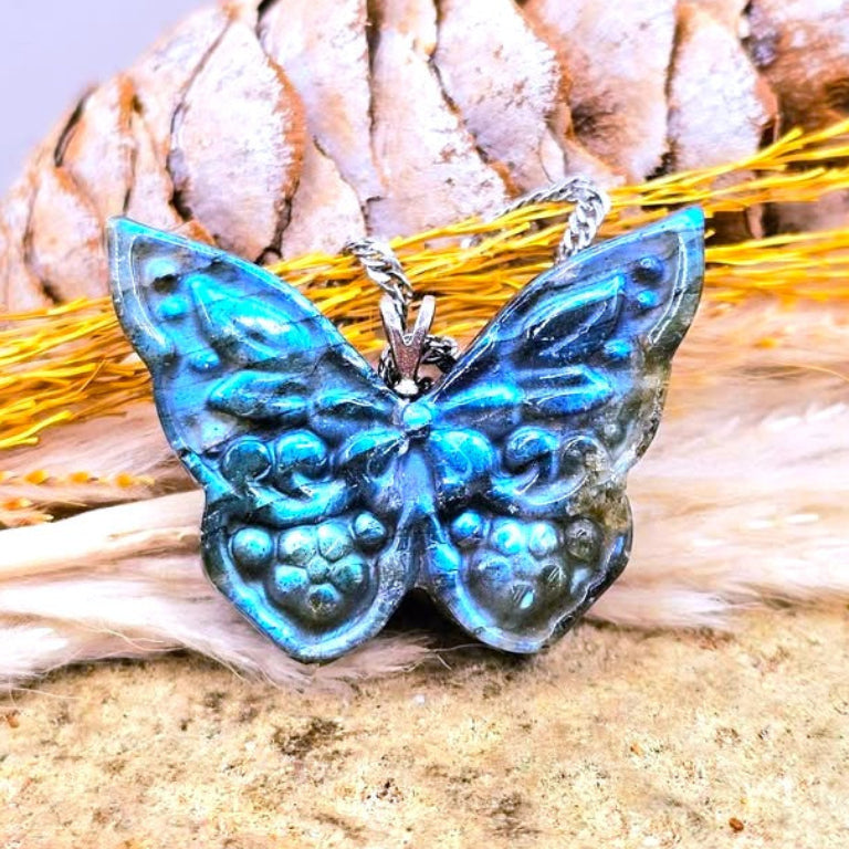 A natural labradorite carved butterfly-shaped pendant with iridescent blue and teal wings, photographed resting on dried grass and sand with a pinecone in the background.