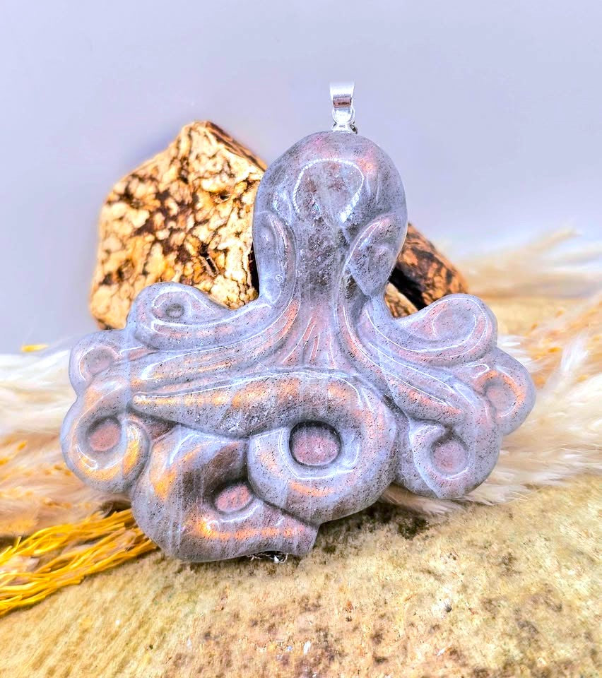 A carved octopus pendant with a blue-green iridescent finish, displayed on a sandy surface with driftwood and a shell in the background.-My Aura Gemstones