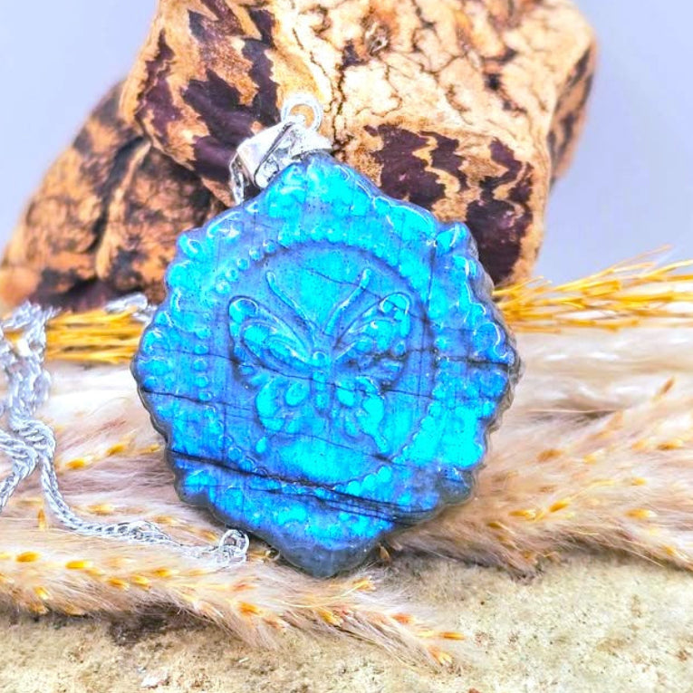 A close-up of a silver-toned pendant necklace with a bright blue flash labradorite carved stone butterfly portrait pendant, styled on a textured surface with a dried pine cone behind it.-My Aura Gemstones