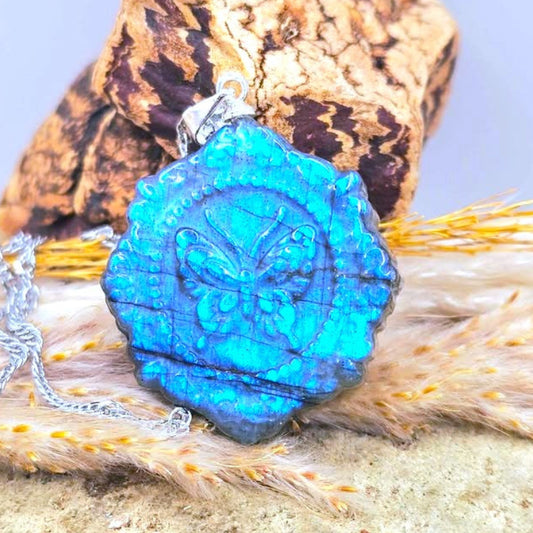 A close-up of a silver-toned pendant necklace with a bright blue flash labradorite carved stone butterfly portrait pendant, styled on a textured surface with a dried pine cone behind it.-My Aura Gemstones