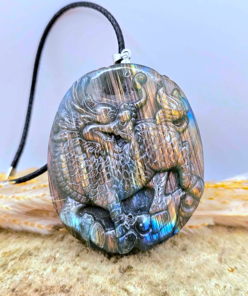 A carved labradorite foo dog pendant necklace with a black cord, displayed on a sandy surface with dried grass and small shells-My Aura Gemstones