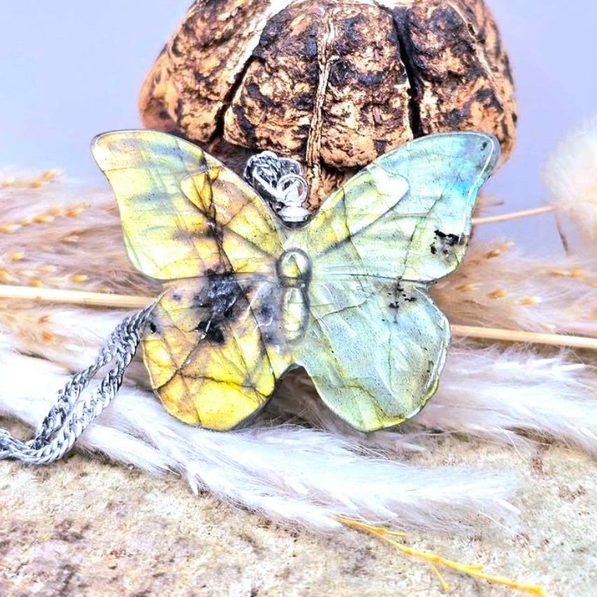 A butterfly-shaped pendant with iridescent blue, green, and yellow wings, resting on dried grass and a textured stone surface with a silver chain.- My Aura Gemstones