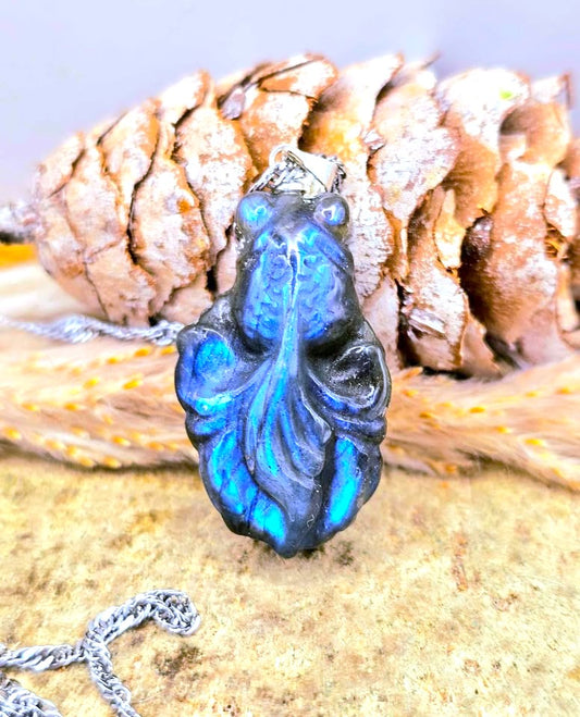 Natural labradorite carved pendant goldfish, featuring vibrant blue and black iridescent coloring, resting on a textured surface in front of a pinecone-My Aura Gemstones