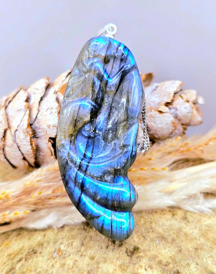 A carved labradorite pendant featuring a mermaid design, hanging from a silver chain against a rustic background of wood and dried grass.-My Aura Gemstones