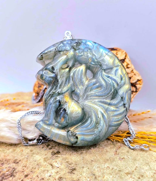 A labradorite carved flashy nine tailed fox sitting on crescent moon pendant featuring a stylized face and flowing hair, with a silver-toned chain. The pendant has a metallic iridescent finish shifting between blue, green, and gold.-My Aura Gemstones
