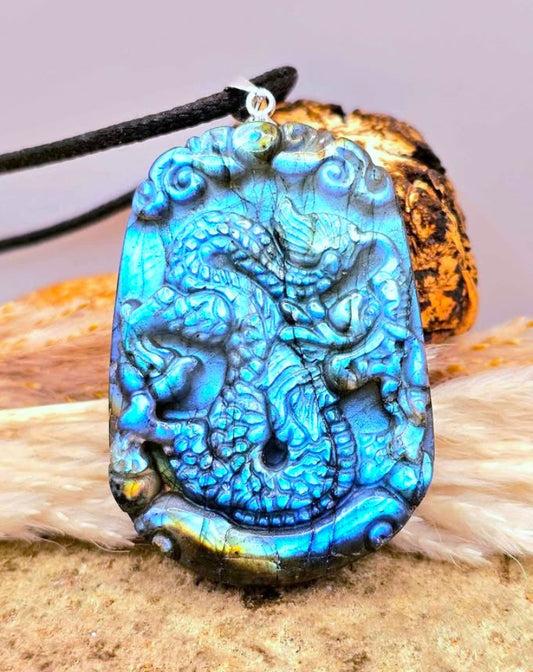 A carved pendant featuring a detailed dragon design in vibrant blue and turquoise labradorite, hanging from a black cord on a rustic background.-My Aura Gemstones