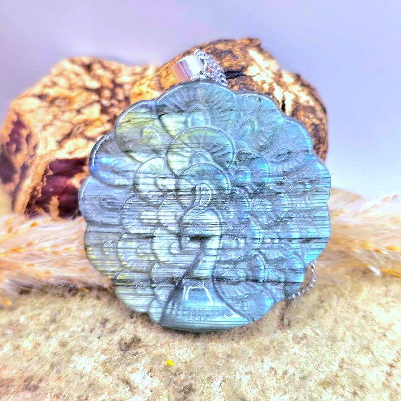 A round labradorite pendant featuring an embossed peacock design, resting on a textured stone surface with a piece of wood in the background.-My Aura Gemstones
