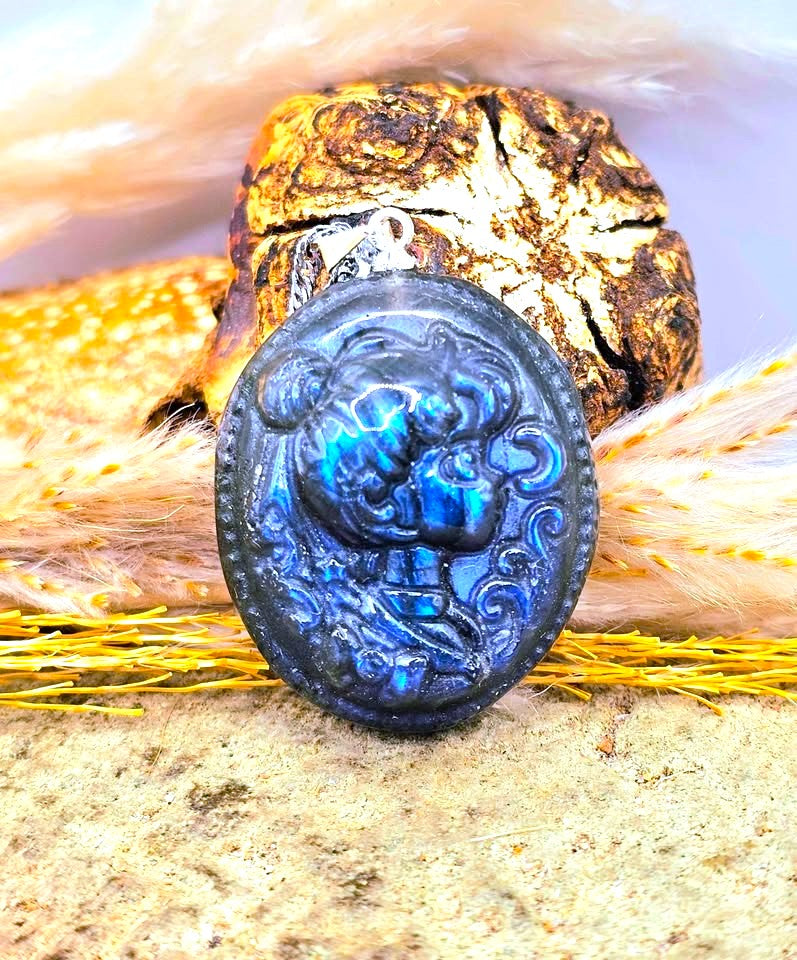 An oval blue labradorite carved sailor pendant or brooch with a raised figure design, displayed on a textured surface with natural props in the background-My Aura Gemstones