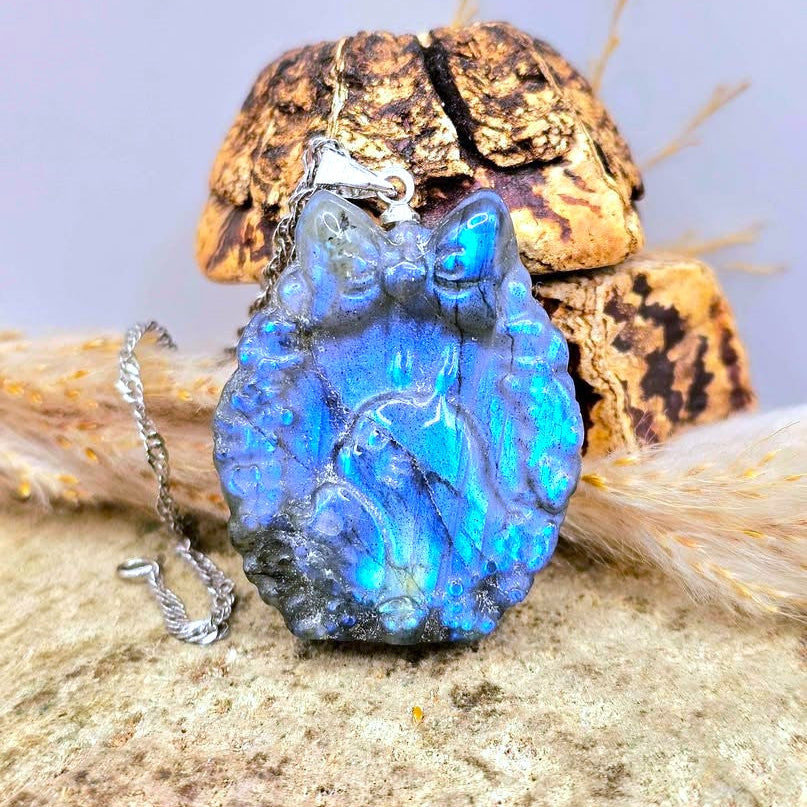 A blue iridescent labradorite carved Totoro portrait pendant necklace with a silver bail and chain, displayed on a textured stone surface with dried grass and bark props.