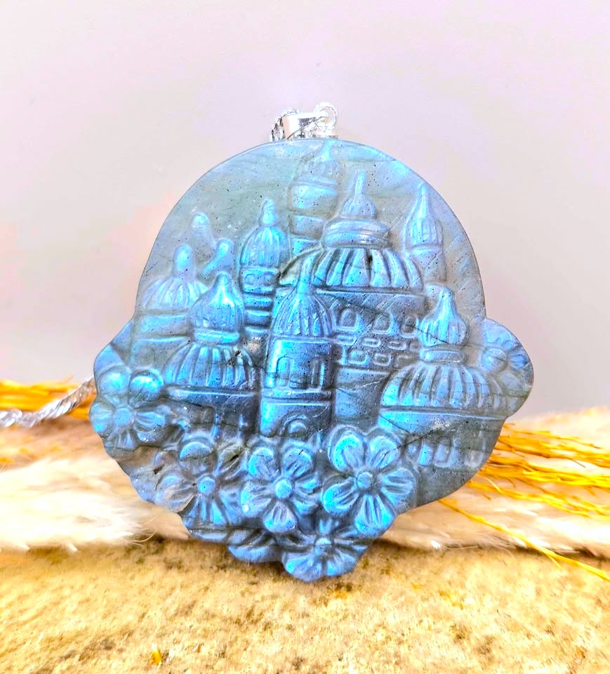 A round blue carved stone pendant featuring a floating castle with domes and flowers, resting on a textured beige surface with soft yellow and white decorative elements in the background-My Aura Gemstones