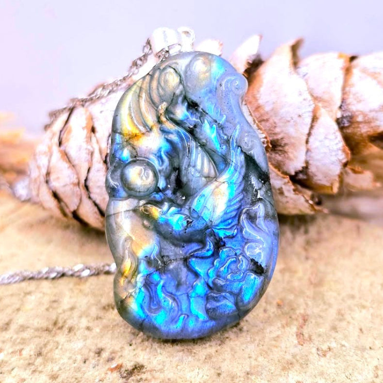 A polished labradorite pendant with an embossed flying phoenix design, resting on a pinecone and textured stone surface. The pendant displays vibrant blue, green, and gold iridescent flashes, suspended from a silver-toned chain.-My Aura Gemstones