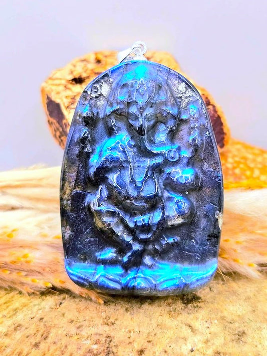 A carved pendant featuring a seated deity with a curved handle, with labradorite with deep blue iridescent flashes with carving of Ganesha on a lotus, displayed on a textured beige surface with dried grass and a blurred background.- My Aura Gemstones