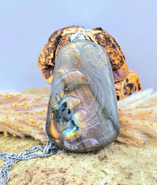 A carved labradorite with two jumping dolphins within waves pendant necklace with a silver chain, displayed on a textured stone surface with dried grass and a decorative object in the background-My Aura Gemstones
