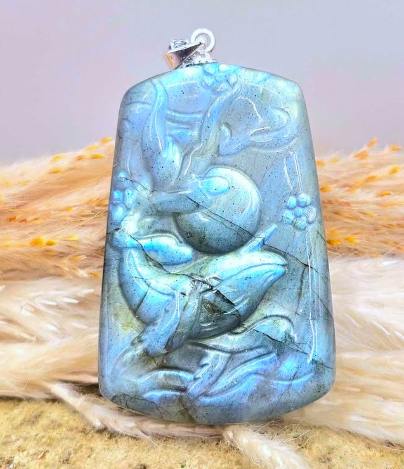 A rectangular labradorite pendant with a carved  a jumping whale, featuring a blue iridescent sheen and a silver bail, displayed on a sandy surface with dried grass.-My Aura Gemstones