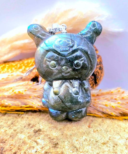 Natural labradorite pendant featuring a Kuromi‑inspired carving with smile and skull detail with silver chain displays on wood slice-My Aura Gemstones