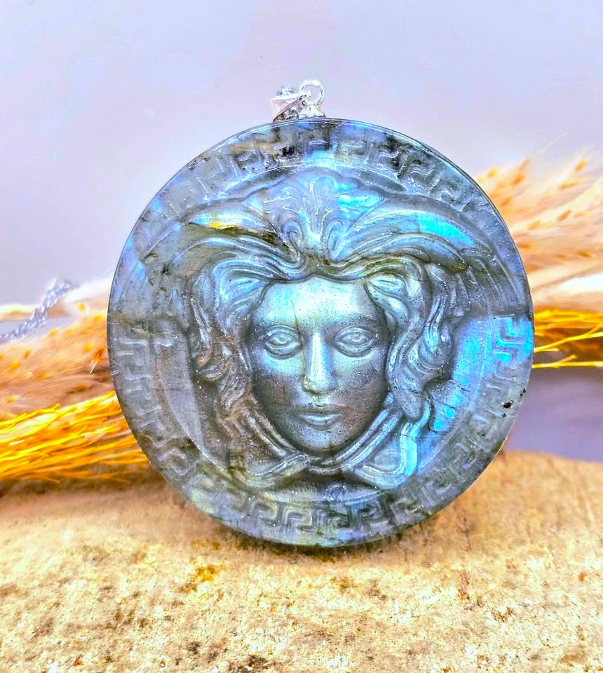A round labradorite pendant necklace featuring a carved Medusa face and Greek key border, resting on a sandy surface with dried grass in the background-My Aura Gemstones
