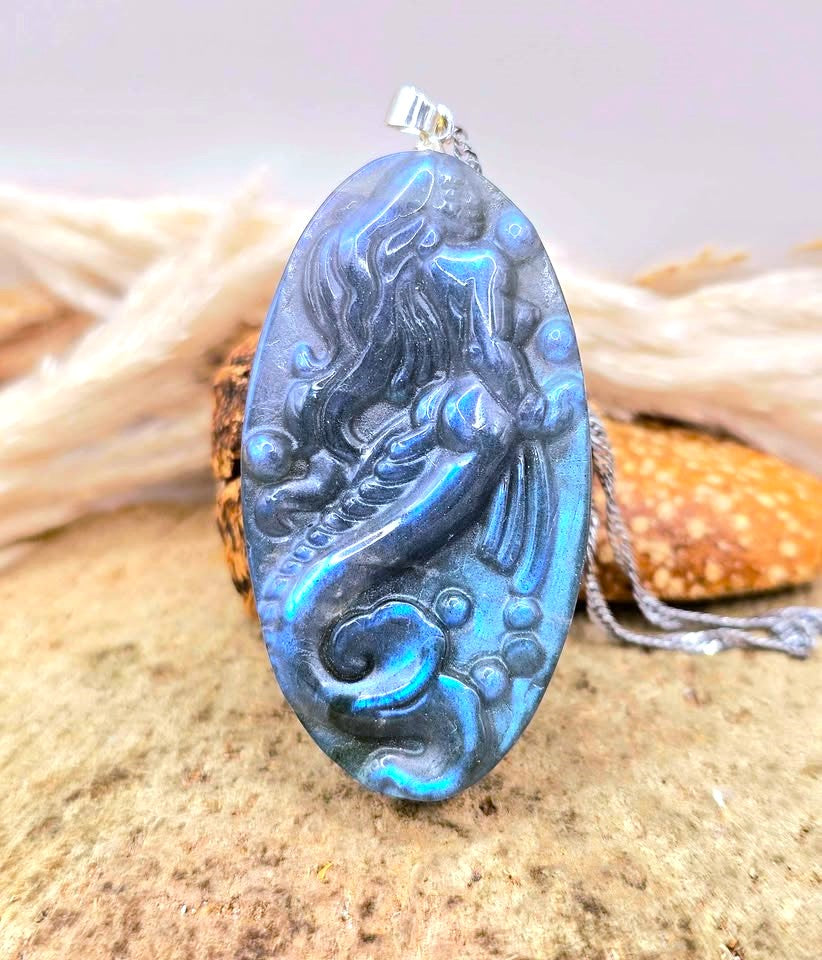 Labradorite carved pendant featuring a mermaid design, suspended from a silver chain, displayed on a sandy surface with dried grass and a small shell in the background-My Aura Gemstones