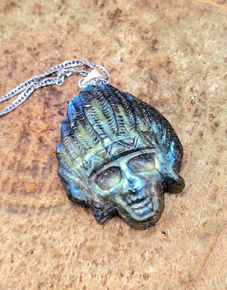 A skull pendant with a blue-green patina and a silver chain, displayed on a textured brown stone surface.