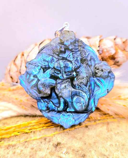 A carved blue labradorite pendant featuring a detailed nine tailed fox design, displayed on a rustic background of dried grass and wood.-My Aura Gemstones