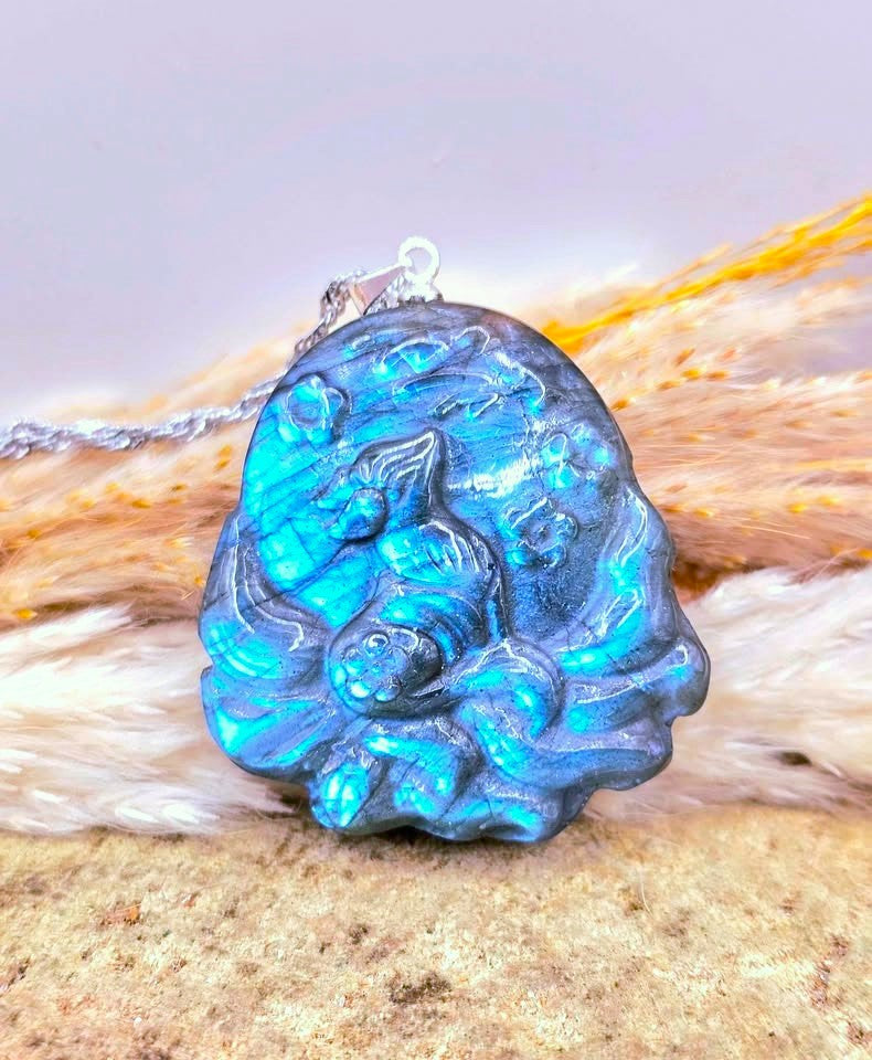 A carved blue labradorite pendant featuring a detailed nine tails fox design, resting on a textured beige surface with a soft white faux fur accent and a silver chain.-My Aura Gemstones