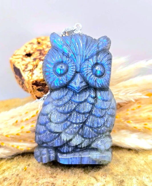 A carved blue owl pendant with iridescent shimmer, displayed on a textured surface with dried grass and a cork-like prop against a soft light gray background.-My Aura Gemstones