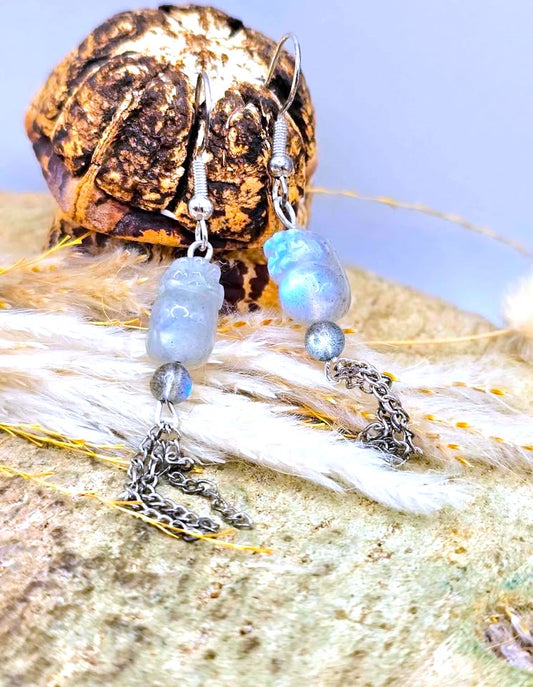 A pair of handmade dangle earrings with labradorite pi xiu earring with 3 chains hanging, silver-toned chain charms, and a small gray bead, displayed on white feathers and a textured brown shell against a pale blue background.-My Aura Gemstones