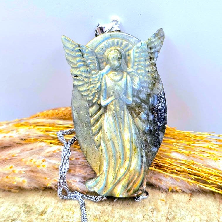 Natural labradorite praying angel pendant necklace with a detailed angel figure and wings, resting on a textured surface with dried grass on a wood slice. -My Aura Gemstones