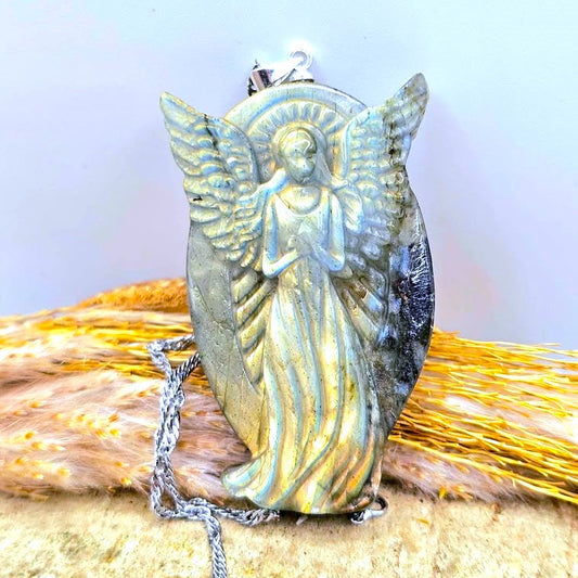 Natural labradorite praying angel pendant necklace with a detailed angel figure and wings, resting on a textured surface with dried grass on a wood slice. -My Aura Gemstones