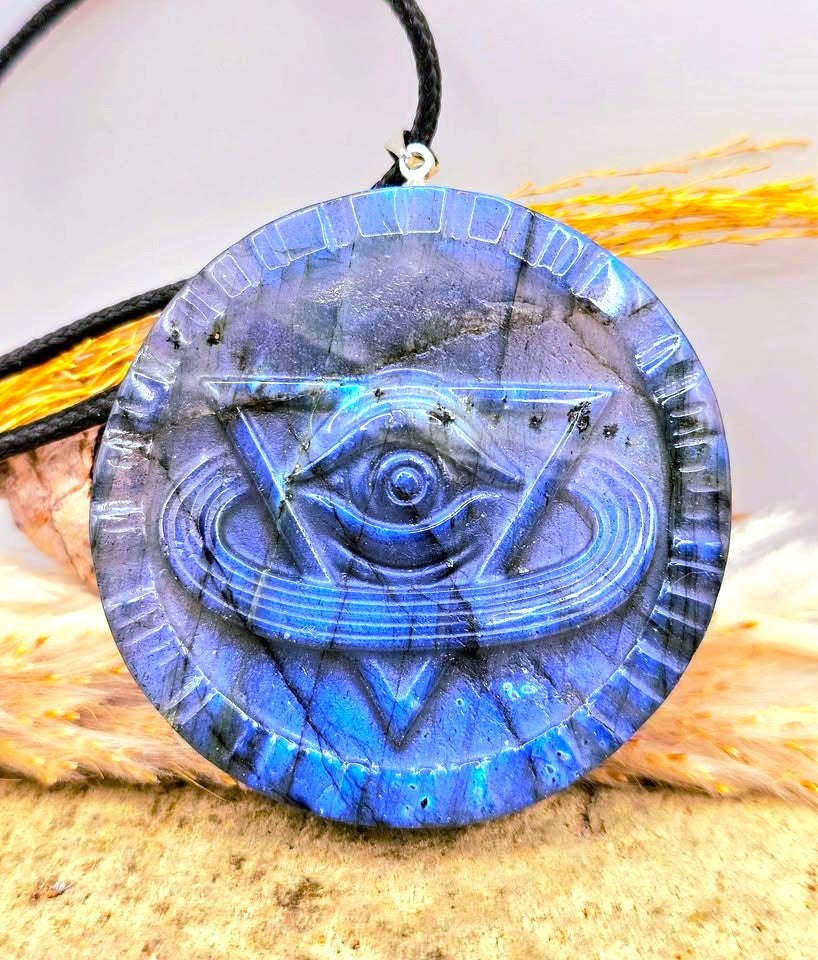 A round blue labradorite pendant necklace featuring an engraved eye of wisdom design, resting on a neutral surface with dried grass-My Aura Gemstones