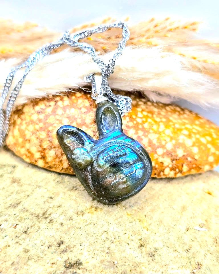 A silver chain necklace with a dark, iridescent green-blue pendant shaped like a rabbit head, resting on a textured sandy surface with a seashell and dried grass in the background.-My Aura Gemstones