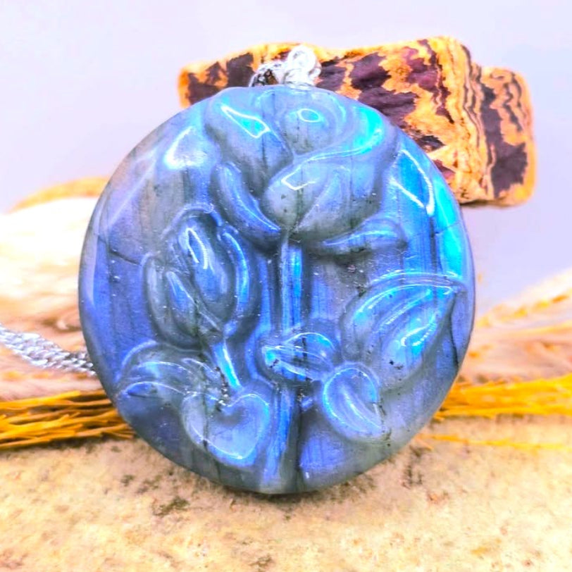 A round labradorite pendant necklace with a carved floral pattern, displaying vibrant blue iridescent flashes, resting on a textured surface with a cork and dried grass-My Aura Gemstones
