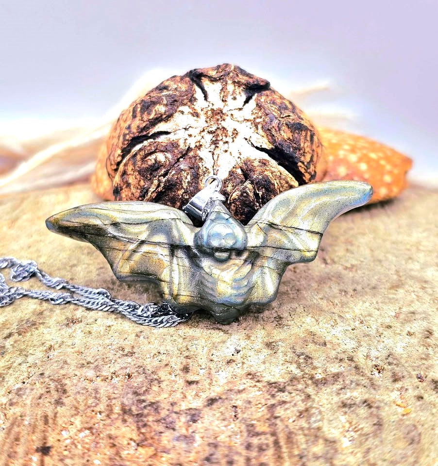 A bat-shaped pendant necklace with a textured, metallic finish, resting on a sandy surface with a dried seed pod in the background.- My Aura Gemstones