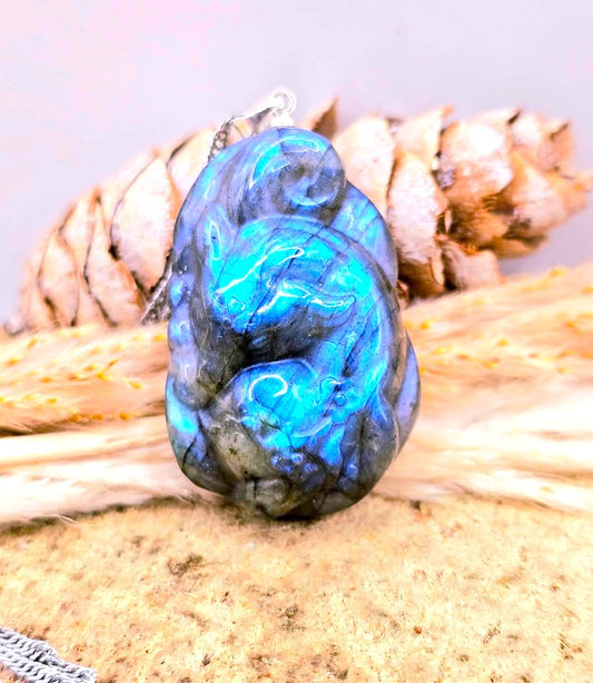 A polished blue labradorite carved sleeping nine tailed fox pendant with swirling iridescent blue, teal, and gray flashes, resting on a textured beige surface with dried grass and pinecones in the background.
