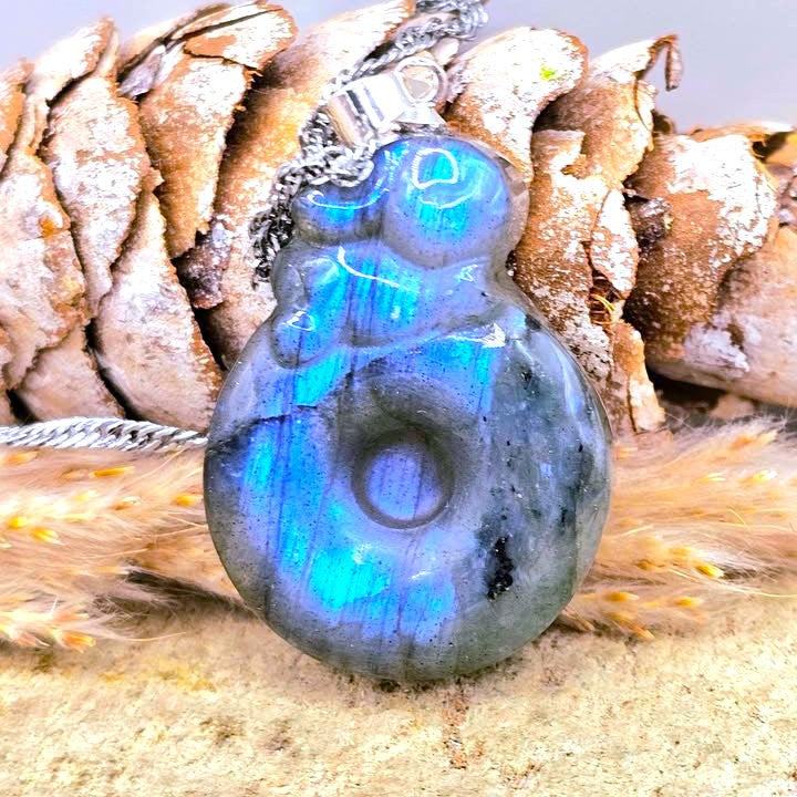 A polished labradorite pendant with carving of rabbit sleeping on a donut necklace with a silver bail, resting on a textured surface with a pine cone and faux fur background.- My Aura Gemstones