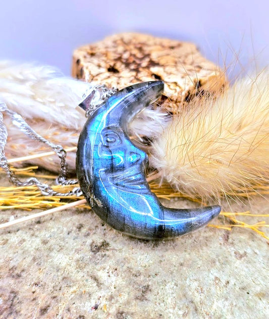 A silver chain necklace with a crescent moon pendant made of iridescent blue labradorite stone, displayed on a textured rock and dried grass.- My Aura Gemstones