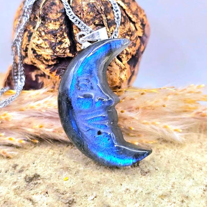 A silver chain necklace with a crescent moon pendant made of iridescent blue labradorite stone, displayed on a textured rock and dried grass.- My Aura Gemstones