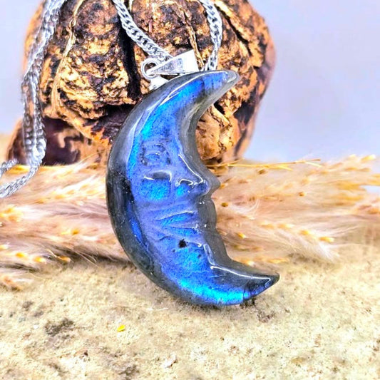 A silver chain necklace with a crescent moon pendant made of iridescent blue labradorite stone, displayed on a textured rock and dried grass.- My Aura Gemstones