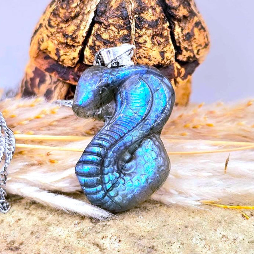 A blue iridescent labradorite snake cobra pendant necklace with a silver bail, displayed on a textured surface with dried grass in the background. - My Aura Gemstones