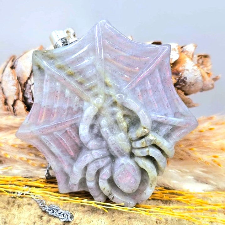 A carved spider pendant or charm made from iridescent pale stone, displayed on a sandy surface with dried grass and a small silver chain nearby.