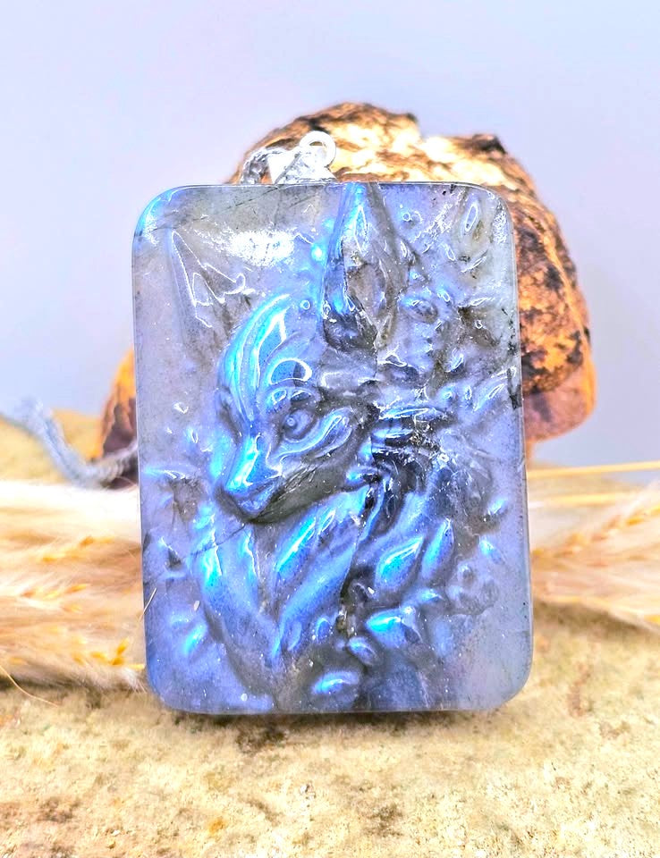 A rectangular pendant featuring a raised wolf head carving, made from a blue iridescent stone with strong blue and purple flashes, displayed on a sandy surface with dried grass and a shell in the background.-My Aura Gemstones