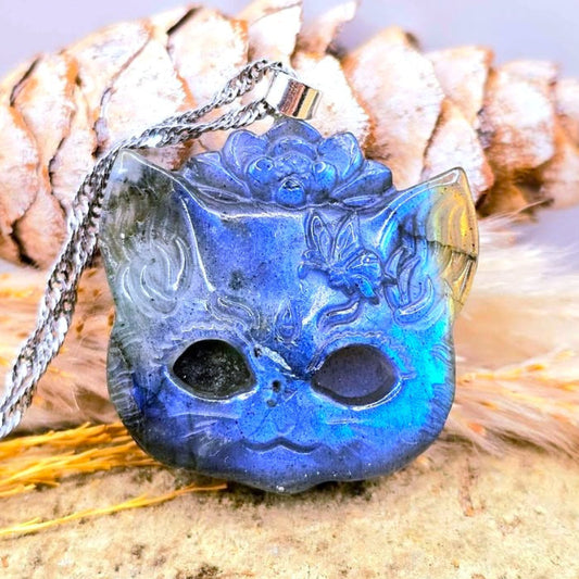A carved blue flash labradorite cat-face mask pendant or ornament with a silver-toned hanging cord, displayed on sand in front of a pinecone and dried grass.-My Aura Gemstones